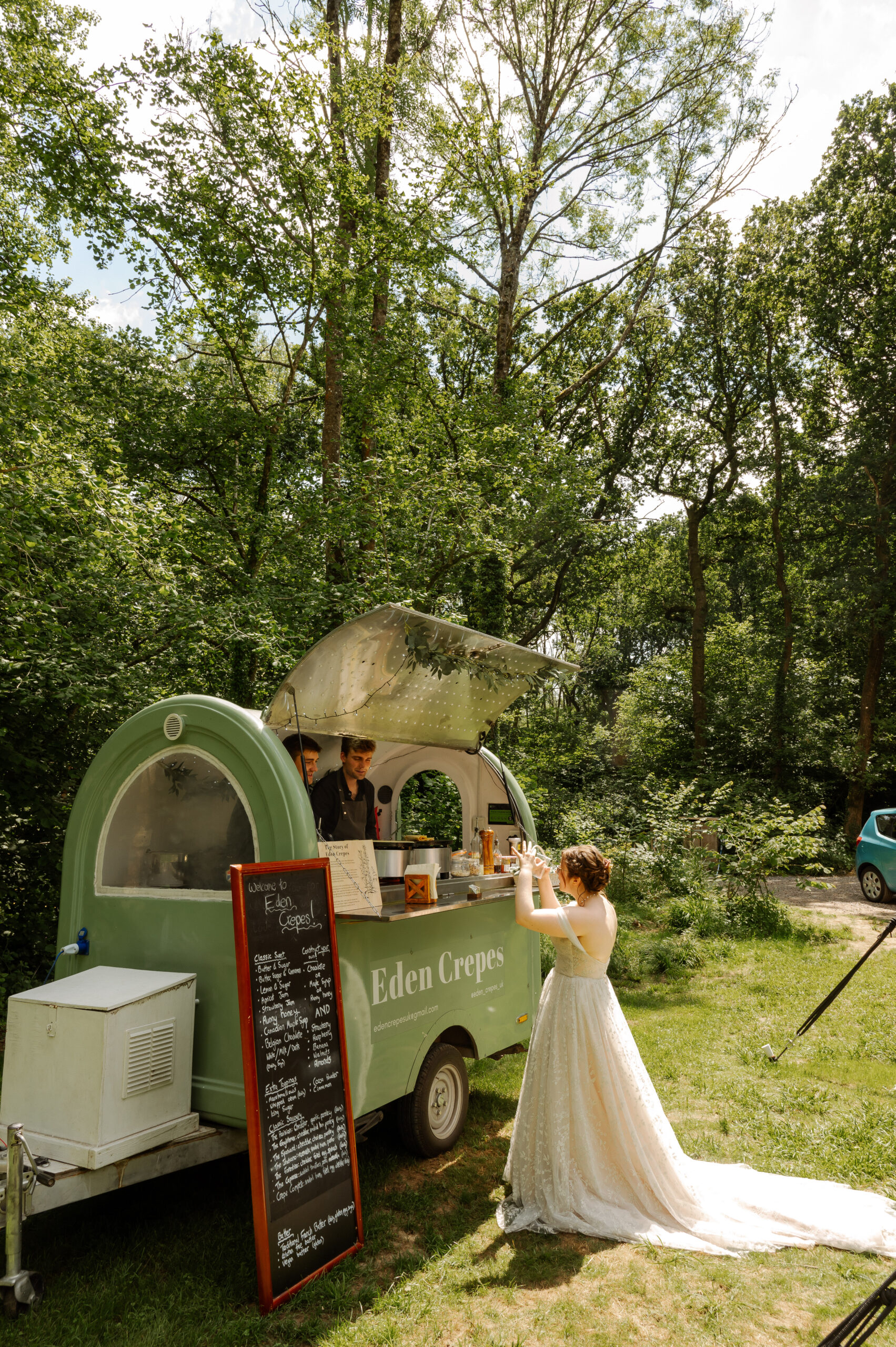 a bride in a white dress stands in front of a crepe stall in a woodland