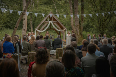 a couple getting married under the oak arbour at the woods at oakley, with guests sitting on benches and bunting and trees surrounding them.