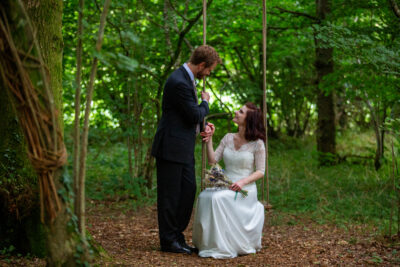 a bride sitting on a swing talking to a groom standing by her in the woods at oakley