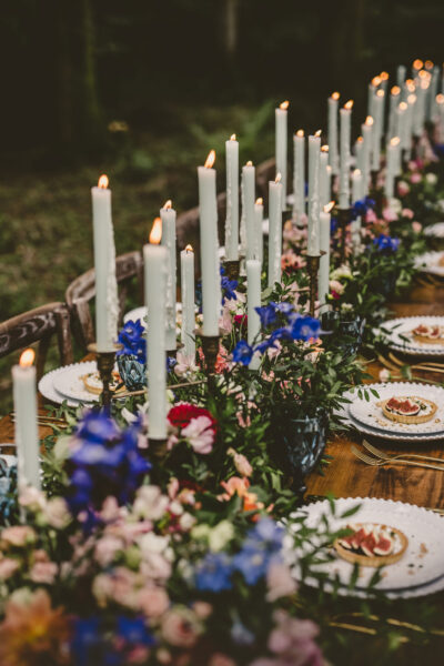 A table decorated with tall white candles, flowers and crockery in a woods.