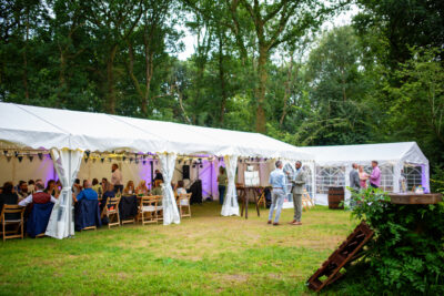 a large marquee in a woodland wedding venue with guests sitting inside and standing outside