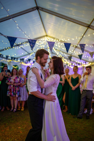 a bride and groom dance in a marquee decorated with fairy lights and bunting, surrounded by their friends