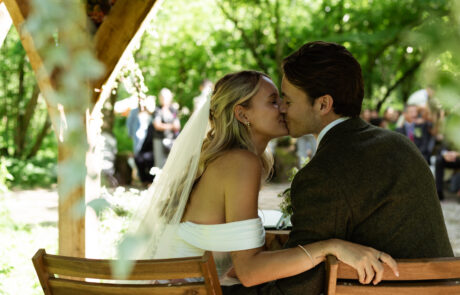 a bride and groom kiss whilst they sit to sign their wedding register at a woodland wedding with their guests in the background