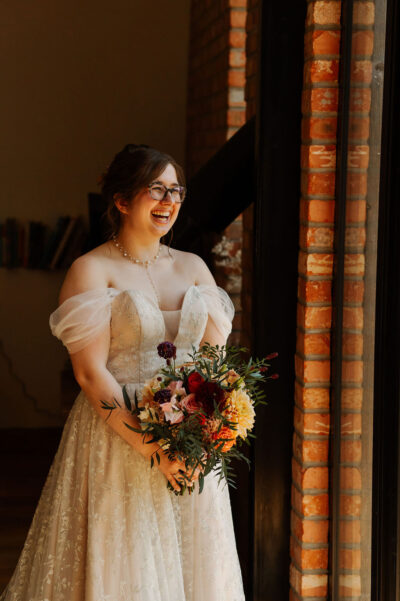 A bride stands looking out of a window and laughing, whilst in her wedding dress and holding a bouquet of flowers.