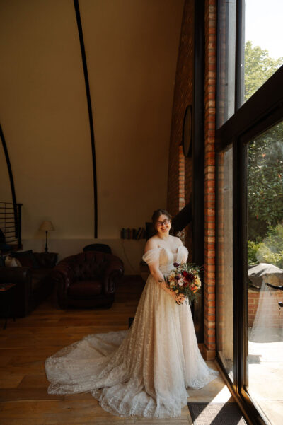 A bride stands in the Nissen Hut, the bridal accommodation at The Woods at Oakley. She is wearing her wedding dress, holding a bouquet of flowers and standing next to a window.