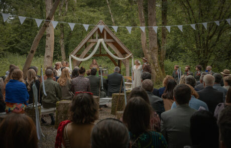 a bride and groom stand with their celebrant in their woodland wedding ceremony, with guests sitting on wooden benches and watching and bunting hanging from the trees