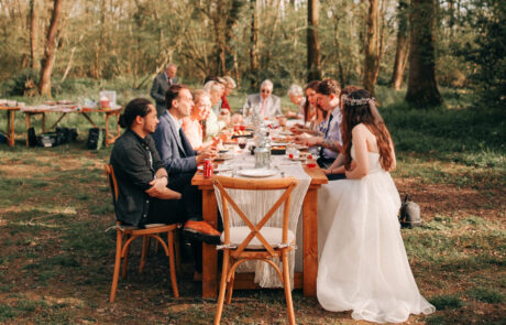 a bride and groom and their guests eat outside in a woodland at a long wooden table