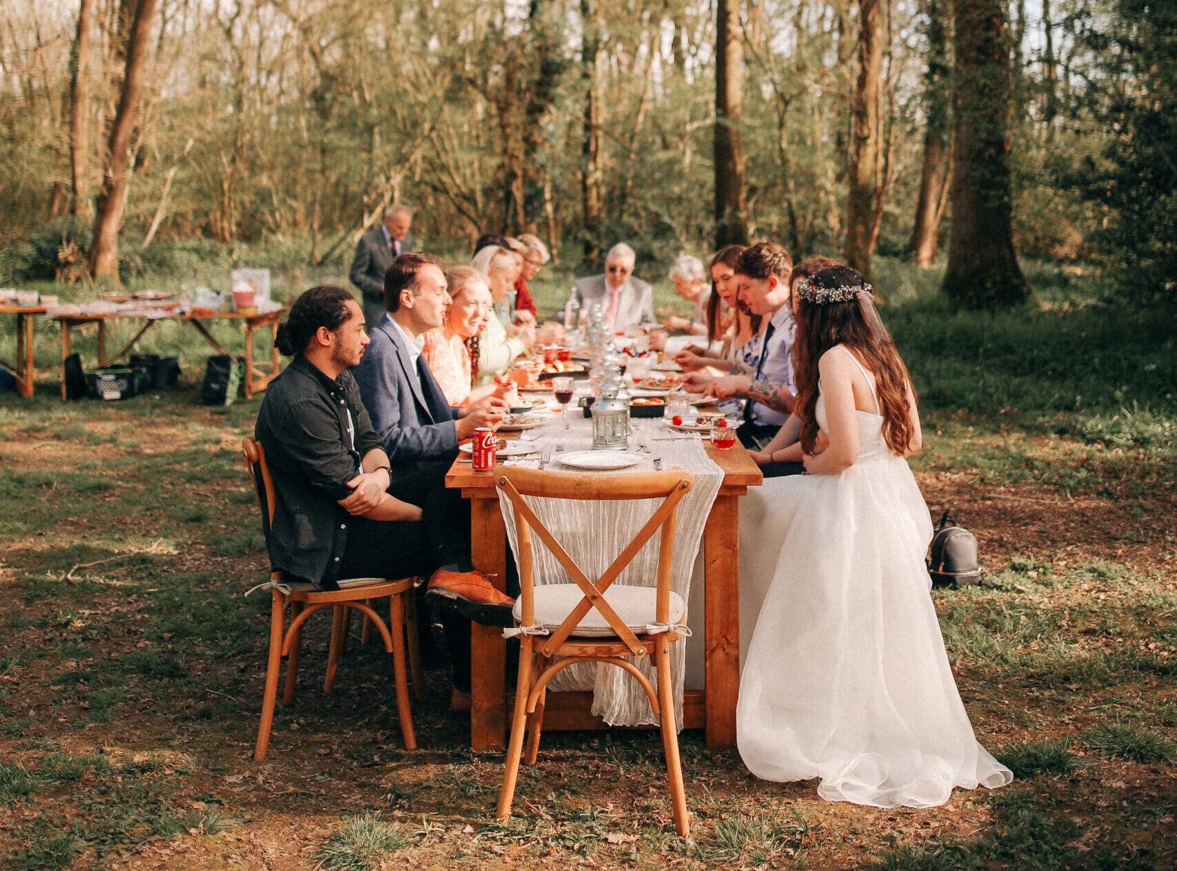 a bride and groom and their guests eat outside in a woodland at a long wooden table