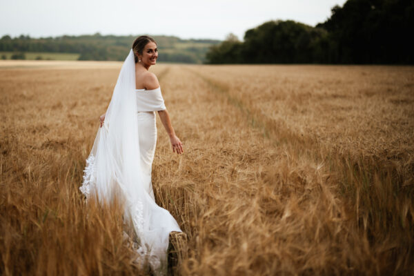 A bride walks through a field in her wedding dress at The Woods at Oakley, looking back and smiling at the camera.