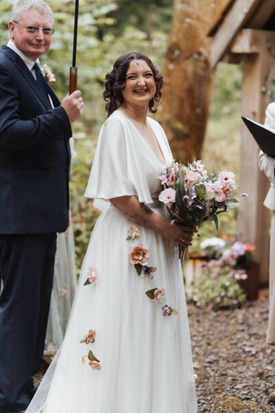 A bride in a white wedding dress adorned with flowers and carrying a bouquet smiles at the camera as she waits to get married at The Woods at Oakley,