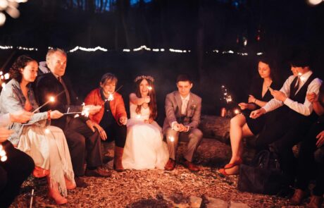 a bride and groom sit with their wedding guests around a fire pit with sparklers in the evening