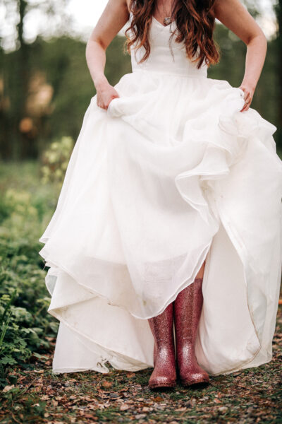 A bride stands in a woodland and holds her wedding dress up to show sparkly pink wellies.