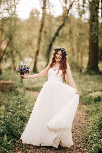 A bride stands in her white wedding dress with a flower crown on, smiling in a woodland and holding her bouquet.