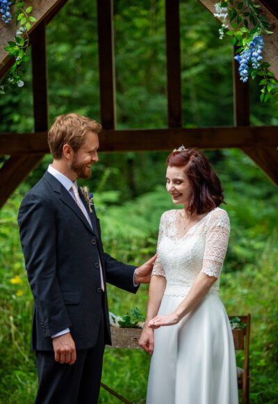 A bride and groom stand next to each other in front of the altar in The Woods at Oakley. They are both laughing and looking at her wedding ring.