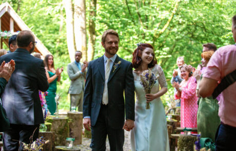 a bride and groom walk back down the aisle together, smiling as their guest applaud in a woodland