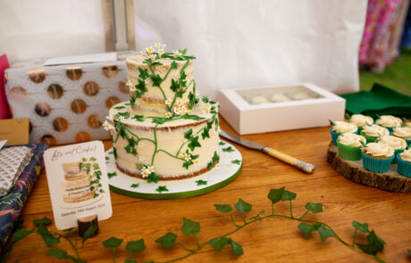 a white wedding cake decorated with iced flowers and ivy sits on a wooden table with presents, cupcakes and ivy on it