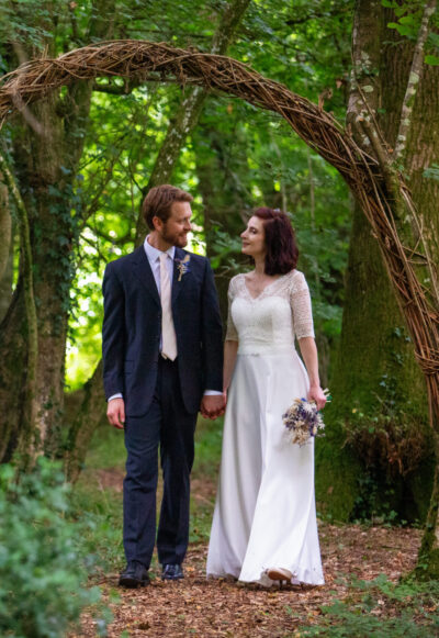 A bride and groom walk through a woodland, looking at each other. 