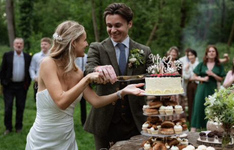 a bride and groom cut their wedding cake outside at a woodland wedding. They smile at each other and their guests watch