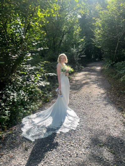 A bride stands in a sunny woodland in her white wedding dress