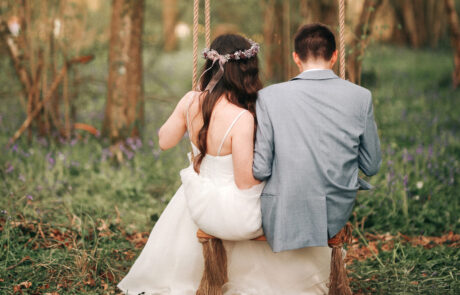 a bride and groom sit on a swing in a woodland with their backs to the camera