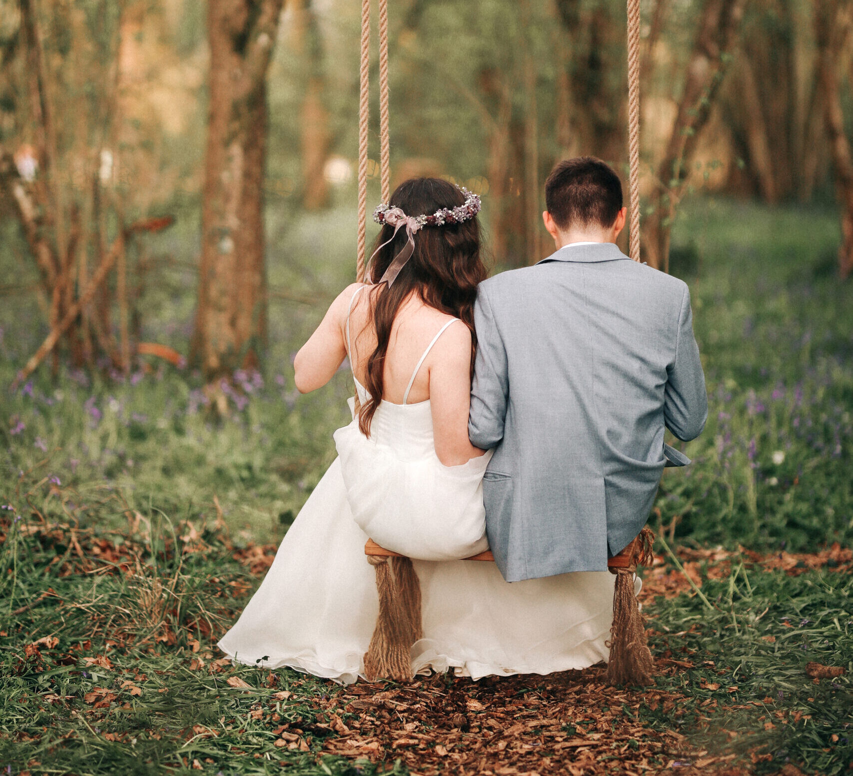 a bride and groom sit on a swing in a woodland with their backs to the camera