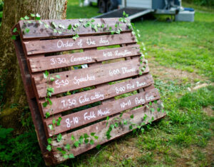 a painted pallet with the order of the day written on it and decorated with ivy leans on a tree, with a horsebox bar in the background