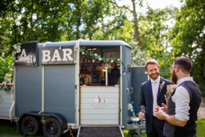 a groom and wedding guest laugh in front of a horsebox bar in a woodland