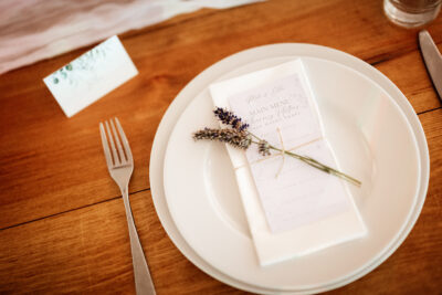 a white plate on a wooden table with a menu tied with a piece of string and adorned with a piece of lavender on top. A knife and fork by the plate and a white place card.