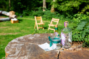 a glass jar with a candle, a glass bottle with fairy lights sit on a stump in a woodland 