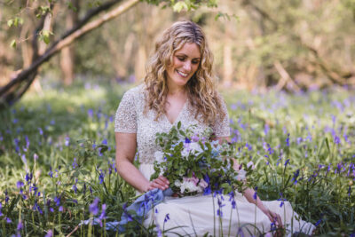 a blonde woman sits in a bluebell woods holding a bouquet of flowers and smiling