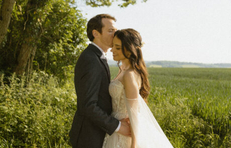 A groom kisses a bride in a green field with blue sky