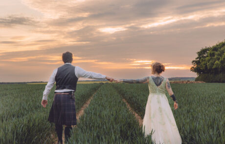 A bride and groom walk hand in hand through a field in a sunset