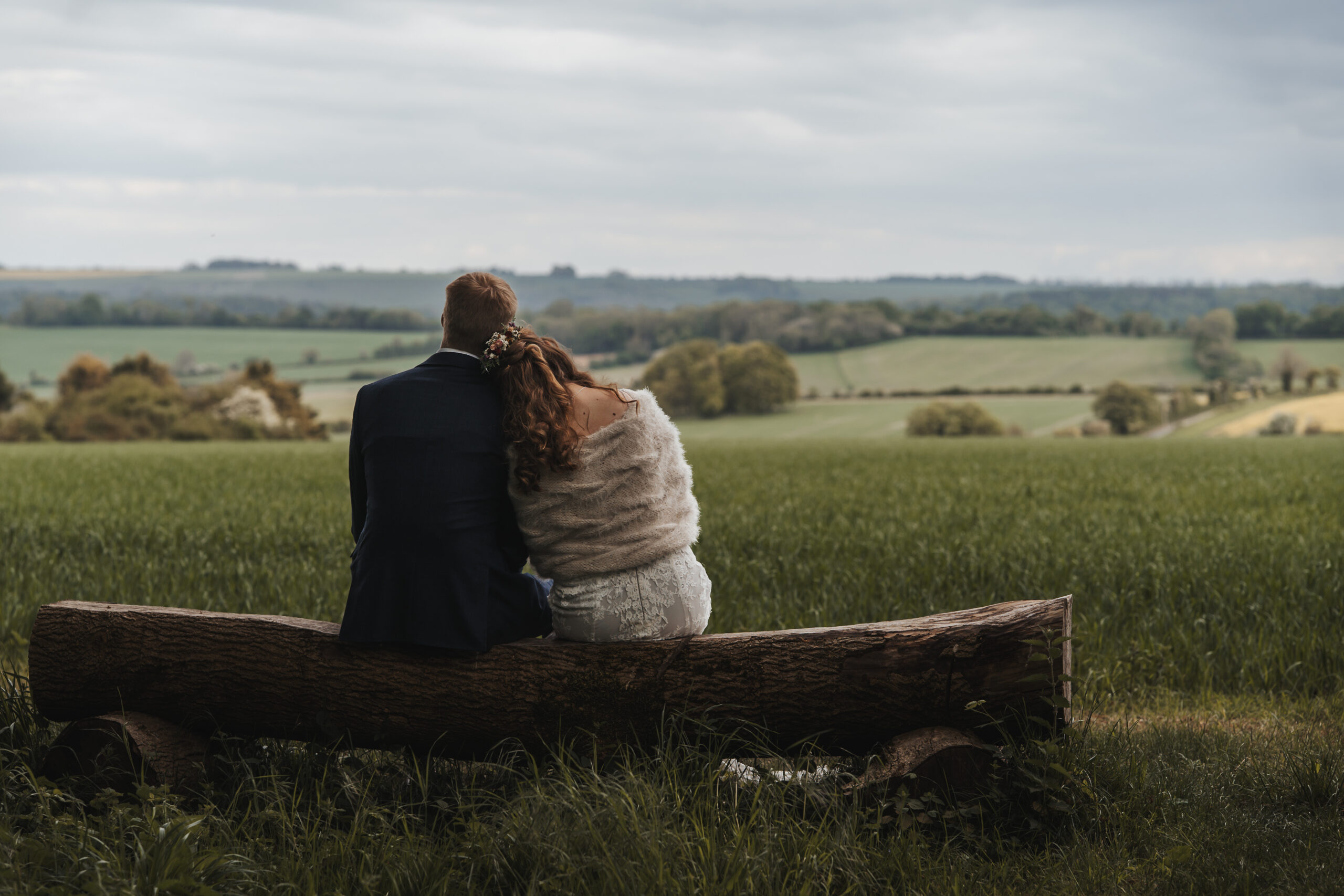 a bride and groom sit on a log bench, looking out at a countryside view