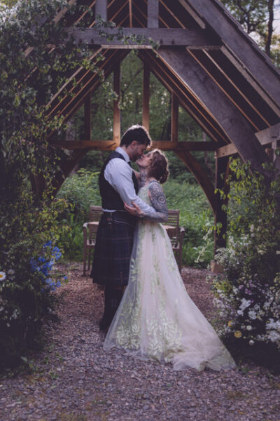 A bride and groom kiss under an oak arbour at a woodland wedding venue