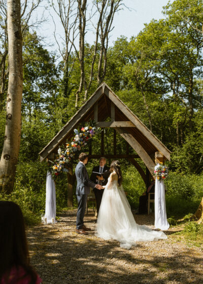 A bride and groom stand under an oak arbour in a woodland holding hands