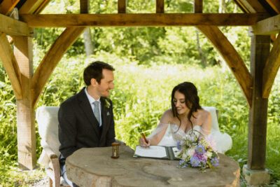 A bride and groom sit at a table signing their marriage certificate in a woodland wedding venue