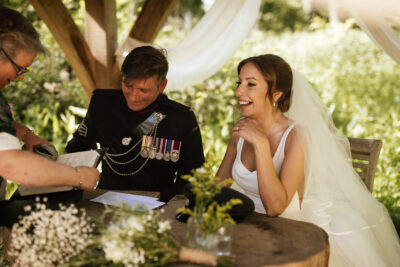 A bride and groom sit at a table in a woods signing their marriage certificate and laughing