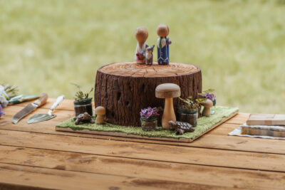 A woodland wedding cake made to look like a tree log with mushrooms around it and a wooden couple on top