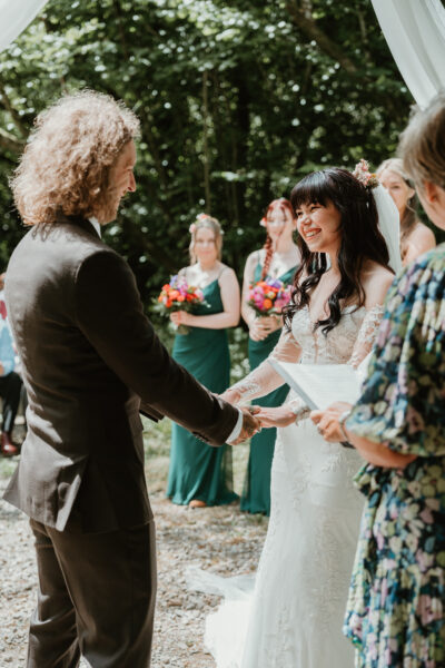 A bride and groom hold hands and laugh at their woodland wedding with guests in the background