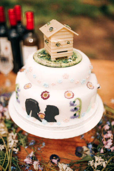 a white wedding cake with a small beehouse on top and surrounded by flowers