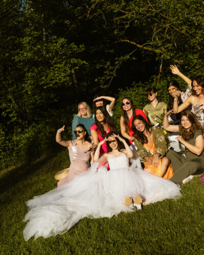 A bride and her bridesmaids sit in a woodland clearing shading their eyes from the sun