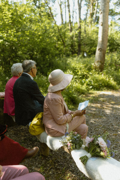 A wedding guest dressed in a hat sitting on a bench in a woodland wedding venue