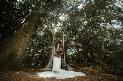 A bride and groom sit on a woodland swing surrounded by trees