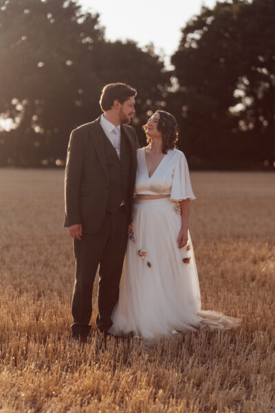 A bride and groom stand in a field during sunset