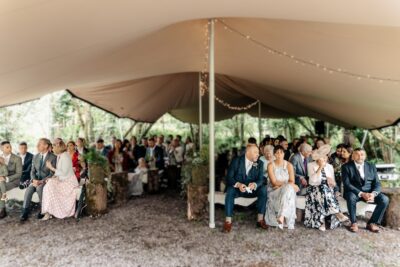 wedding guests sit under a stretch canvas on benches in a woodland