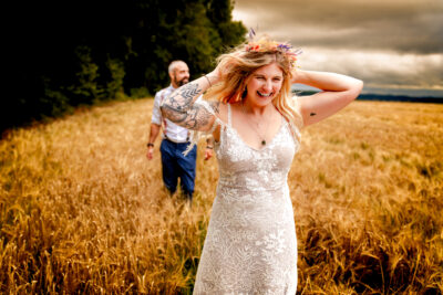 a bride and groom walk through a field in stormy weather laughing