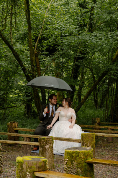 a bride and groom sit on a bench under an umbrella in a woodland