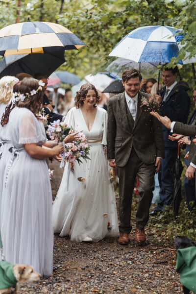 a bride and groom walk through a woodland with their guests holding umbrellas